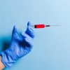 Close-up of a gloved hand holding a syringe with red fluid against a blue background, symbolizing healthcare and medicine.